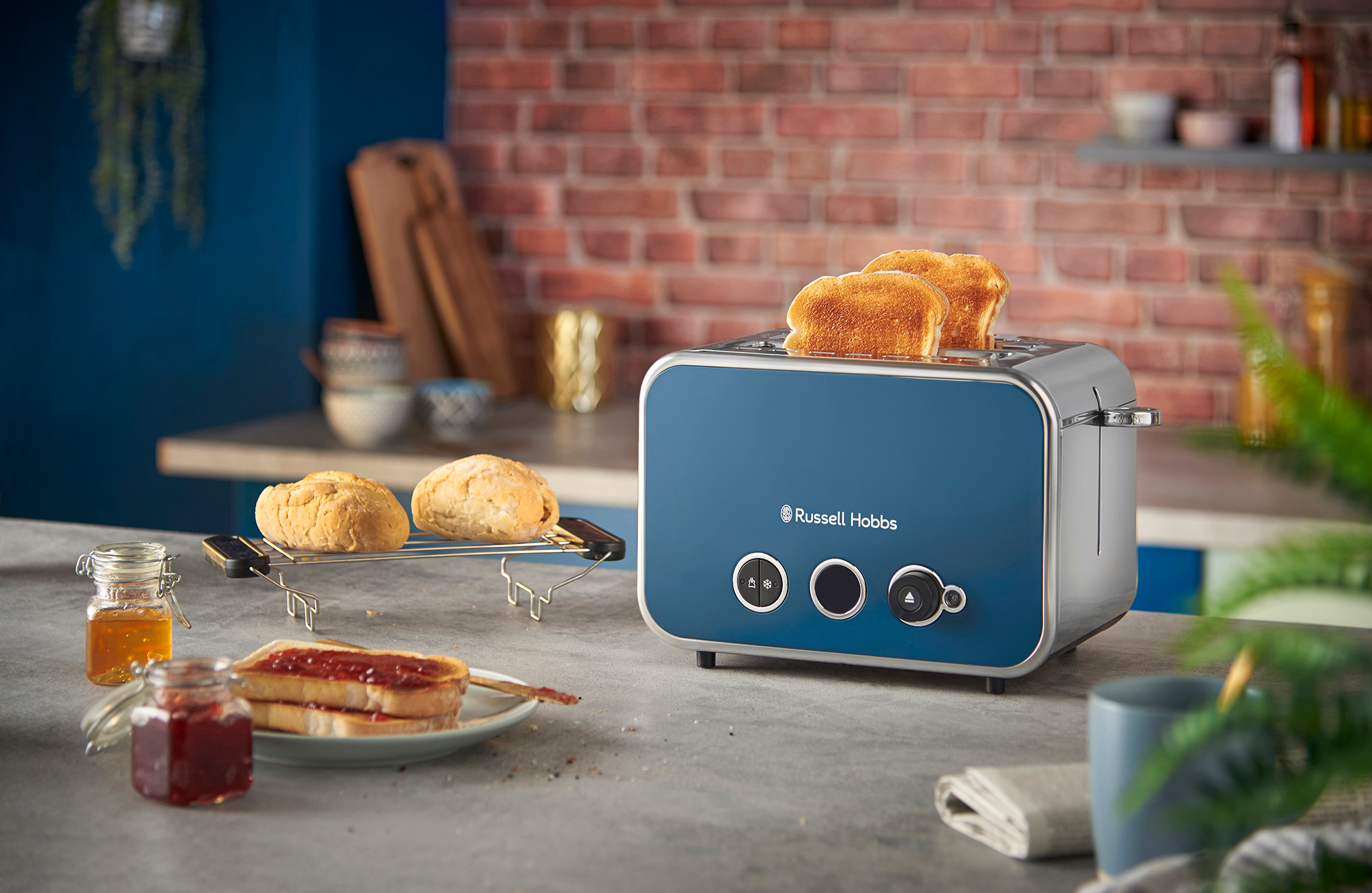 Kék Russell Hobbs kenyérpirító pirítóssal, zsemlékkel, lekvárral és mézzel a pulton.