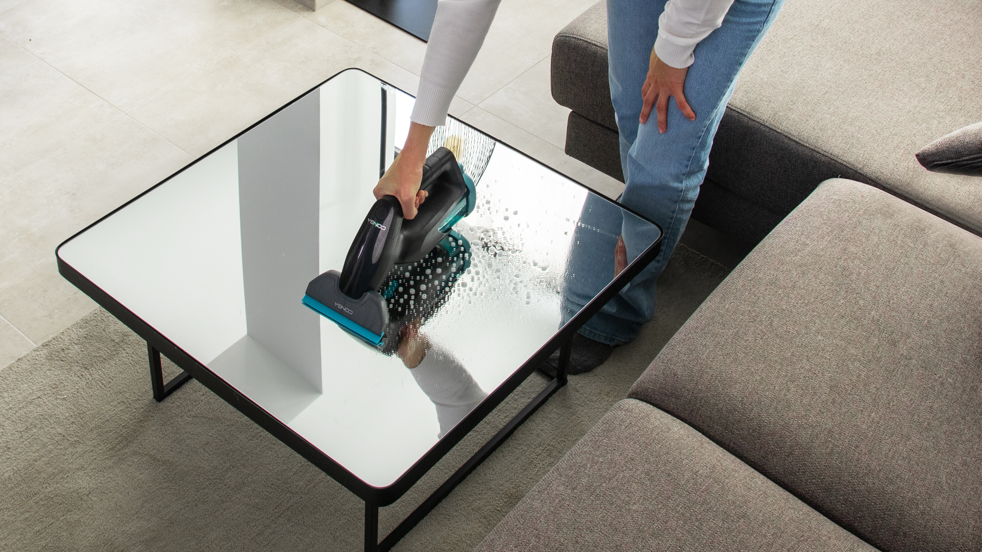 Una mujer limpia una mesa de cristal con una herramienta de limpieza negra, reflejada en el cristal.