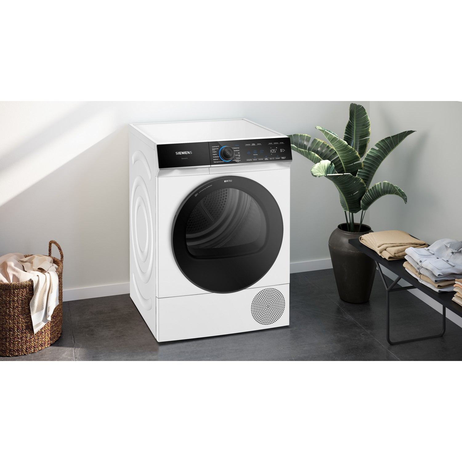 Witte droger met zwart bedieningspaneel en wasmand en plant in de scène.