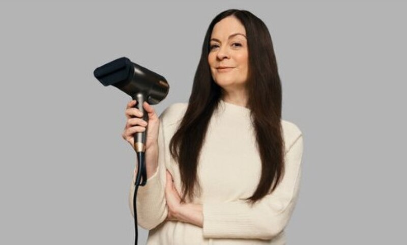 Kobieta trzymająca czarną suszarkę do włosów. Długie brązowe włosy, biały sweter, szare tło.