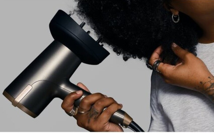Osoba używająca czarno-złotej suszarki do kręconych czarnych włosów. Na dłoni są pierścienie.