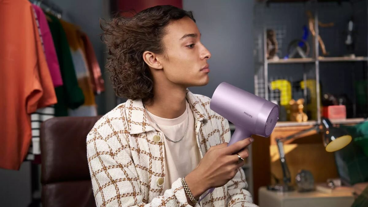 Mężczyzna trzymający fioletową suszarkę do włosów Philips, w koszuli we wzory i z kręconymi włosami.
