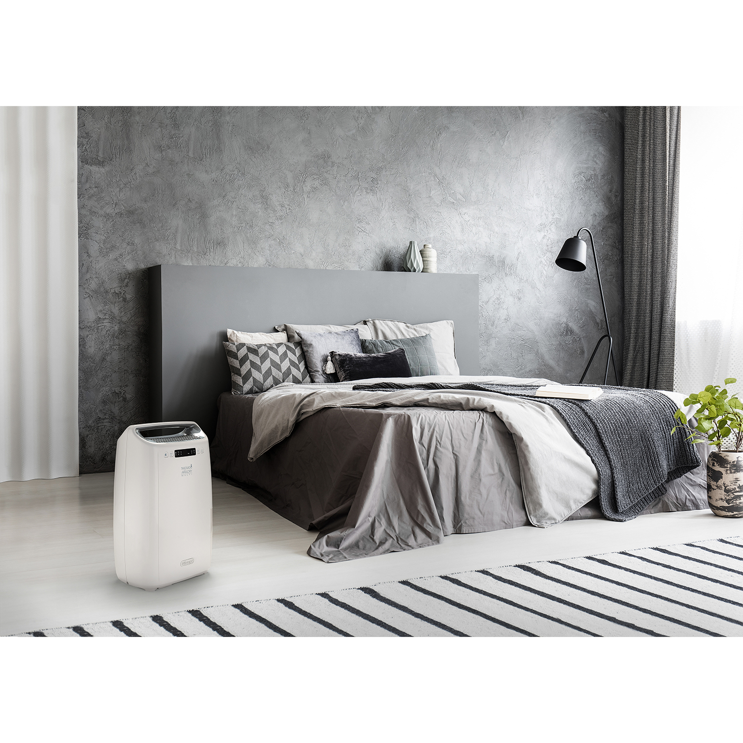 Un purificatore d'aria bianco accanto a un letto. Testata e parete grigie, un tappeto a strisce bianche e nere.