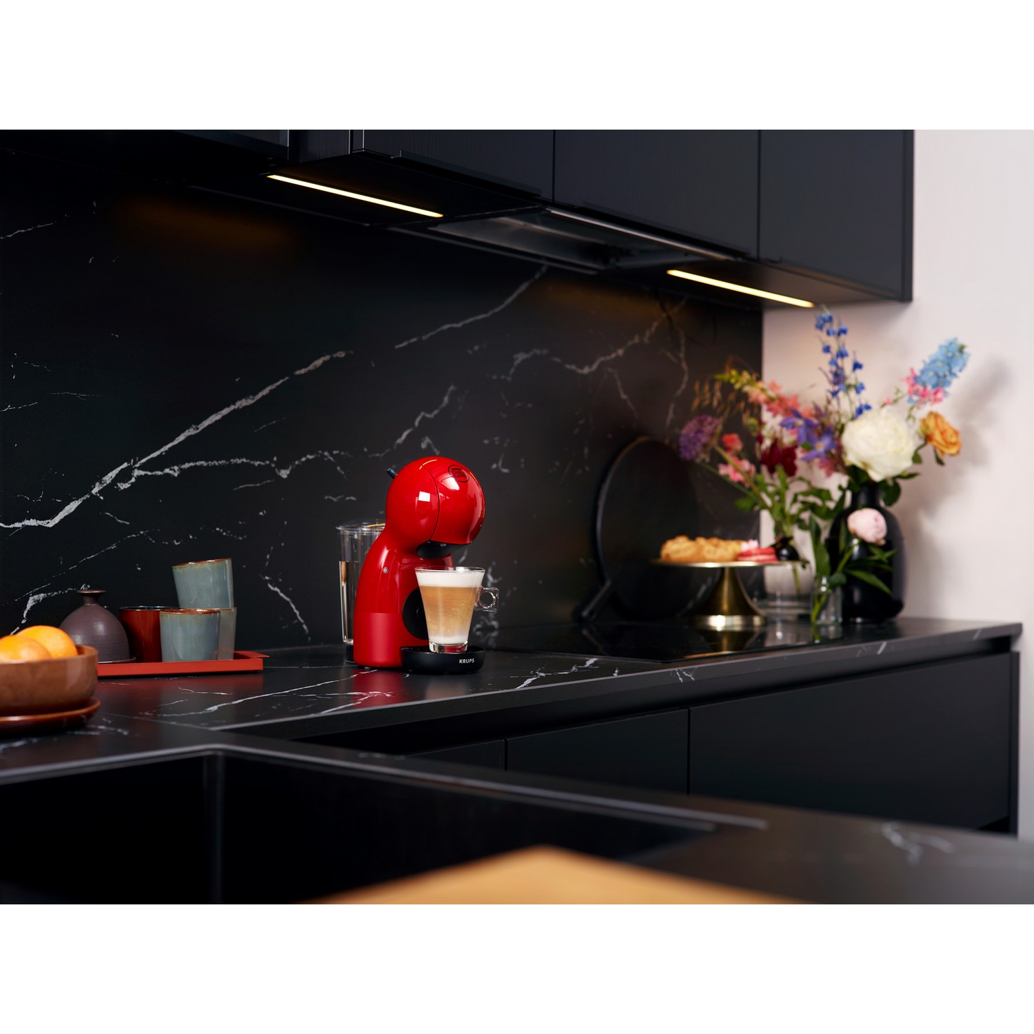 Rode koffiemachine, glas, kopje, bloemen en eten op zwart marmeren aanrecht.