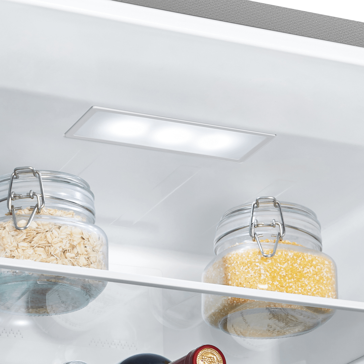 Dentro de un refrigerador, dos frascos de vidrio que contienen avena y granos amarillos con una luz arriba.