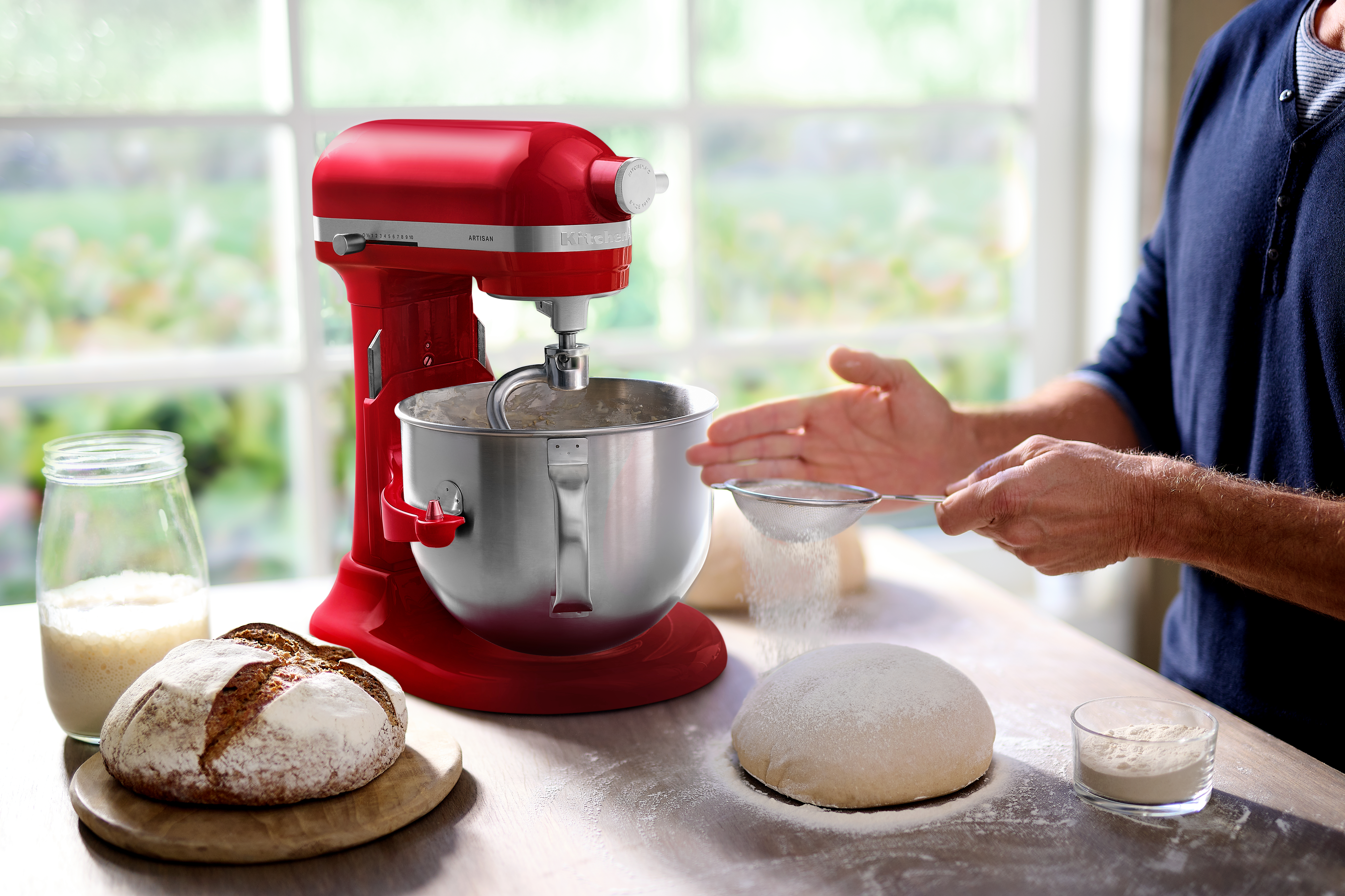 Ein roter Küchenmixer mit silberner Schüssel, Brot und Teig. Eine Person siebt Mehl.