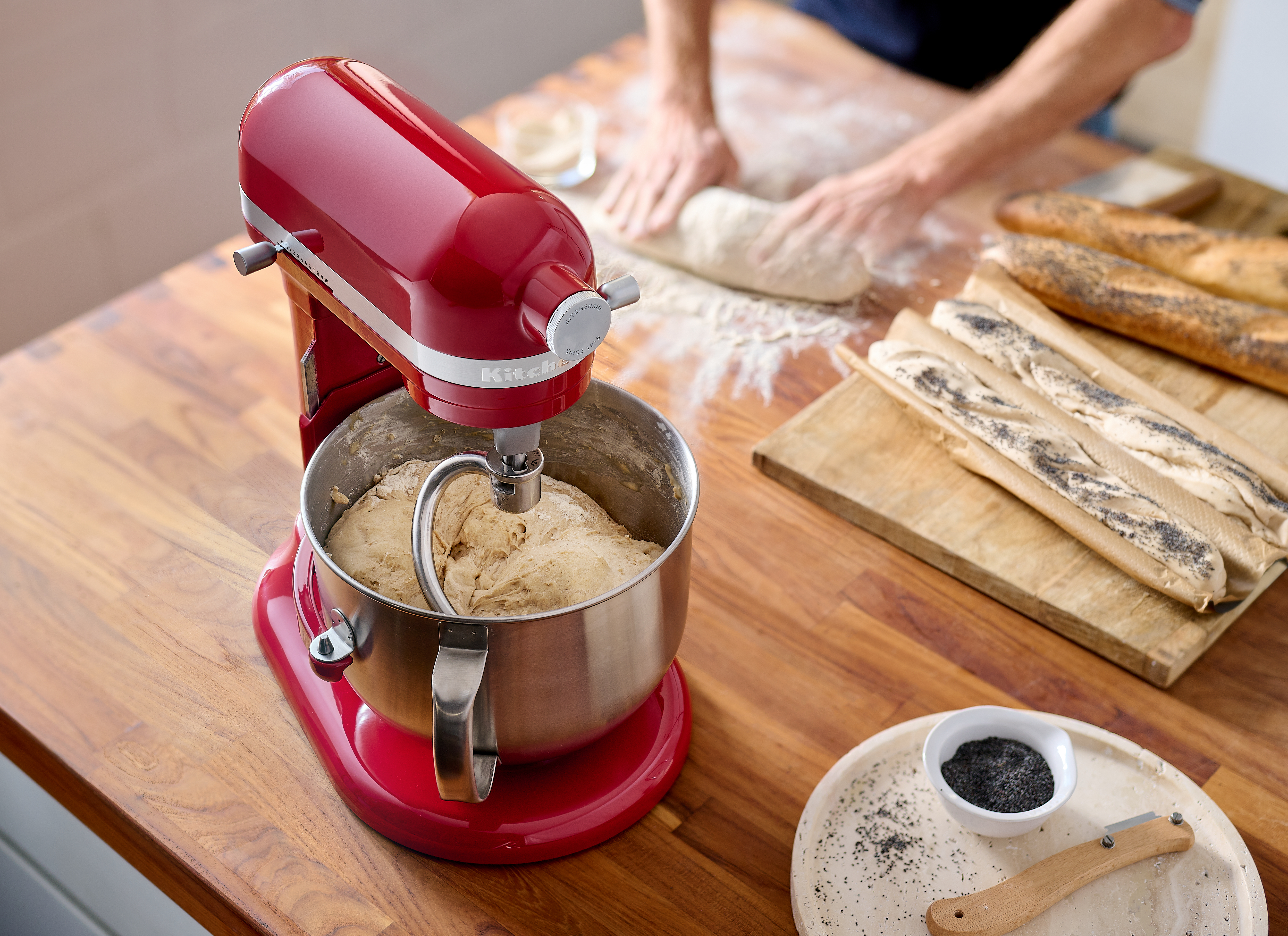 Ein roter Küchenmixer mit Teig in der Schüssel, Brot und Zutaten auf einem Holztisch.