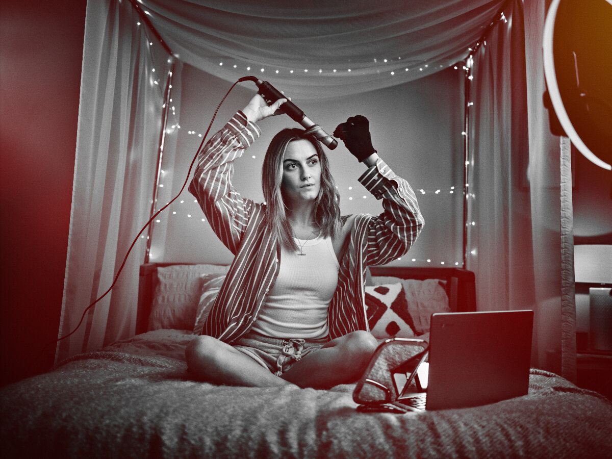 Kobieta siedzi na łóżku, trzyma lokówkę. Światła i laptop w tle.