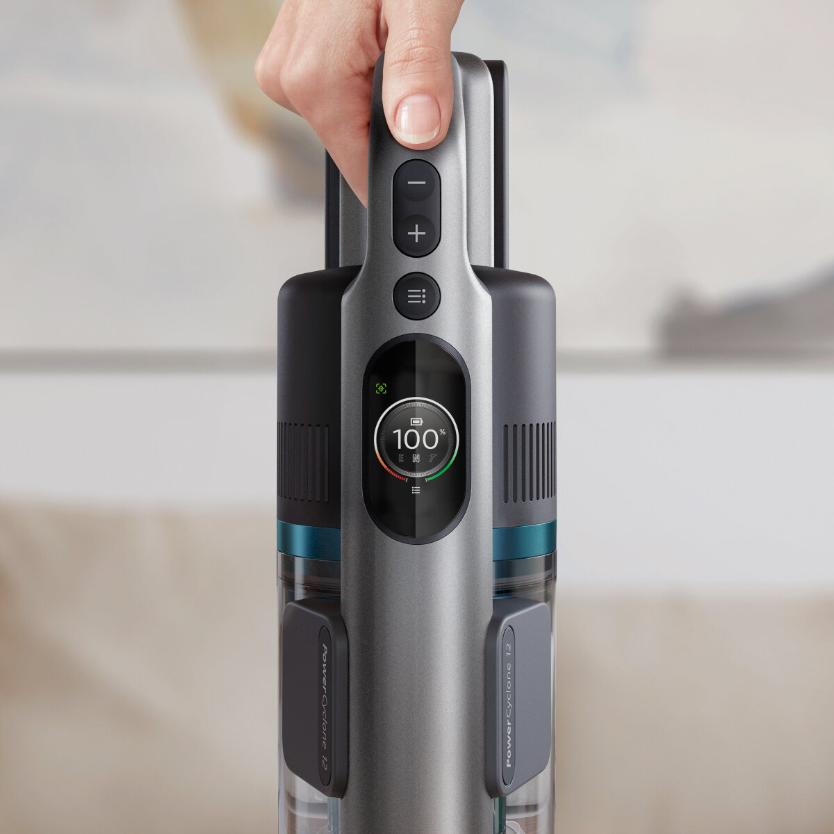 Ręka trzyma szary uchwyt odkurzacza. Ekran pokazuje poziom naładowania baterii.
