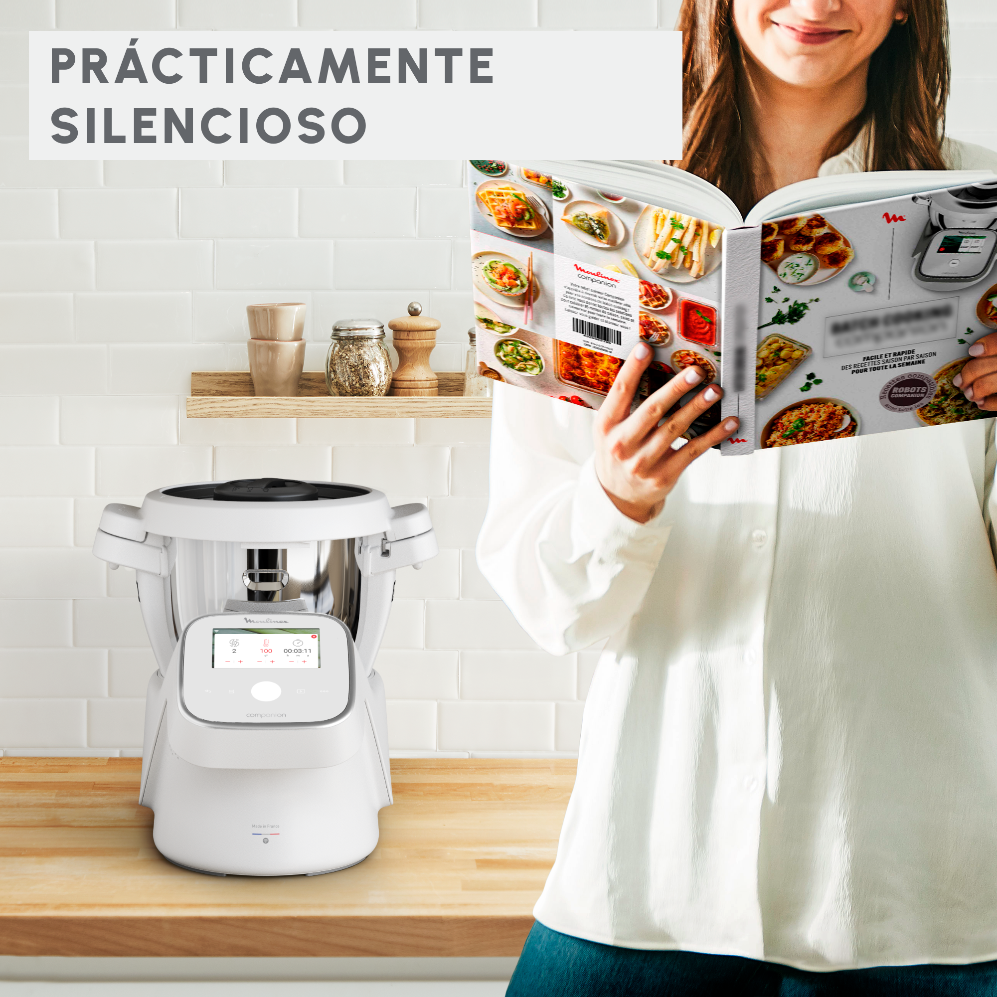Una mujer lee un libro de cocina frente a un electrodoméstico blanco en una pared de azulejos blancos.
