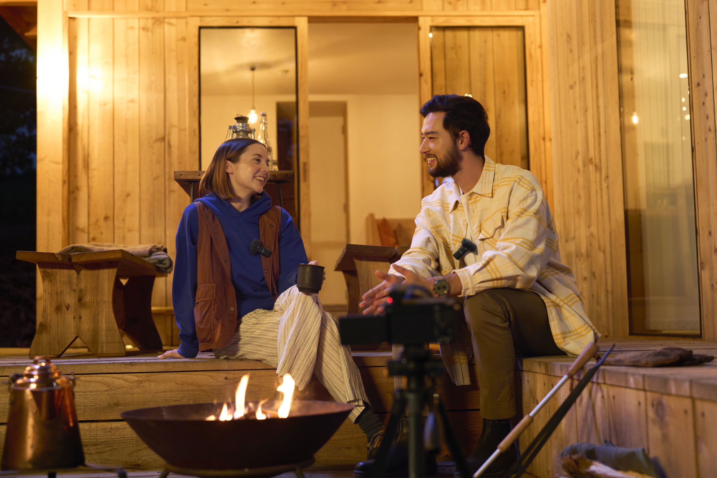 Zwei Personen unterhalten sich auf einer Holzveranda mit Lagerfeuer und Kameraausrüstung.