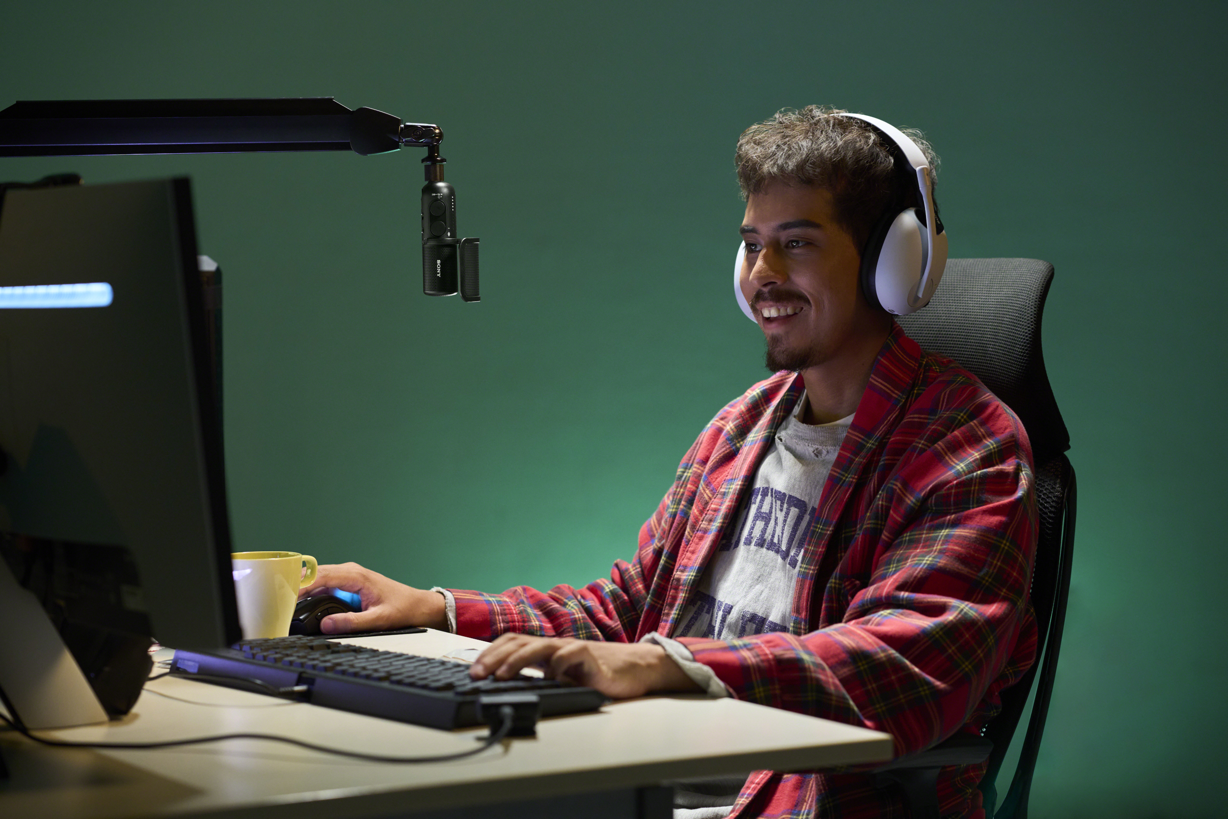 Mann in rotem karierten Hemd und Kopfhörern lächelt beim Spielen am Computer. Eine gelbe Tasse in der Nähe.