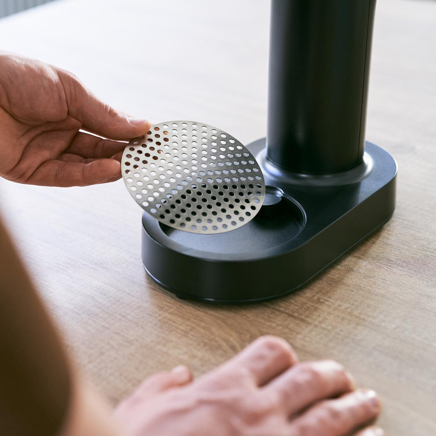 Osoba trzyma perforowany filtr metalowy. Filtr jest czarny i okrągły. Czarny przedmiot na drewnie.