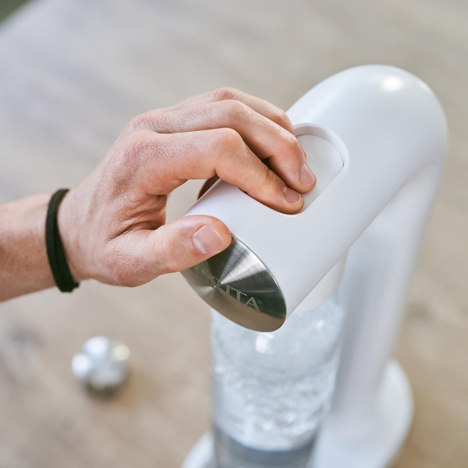 Dłoń naciska przycisk na białym ekspresie do sody. Poniżej butelka.