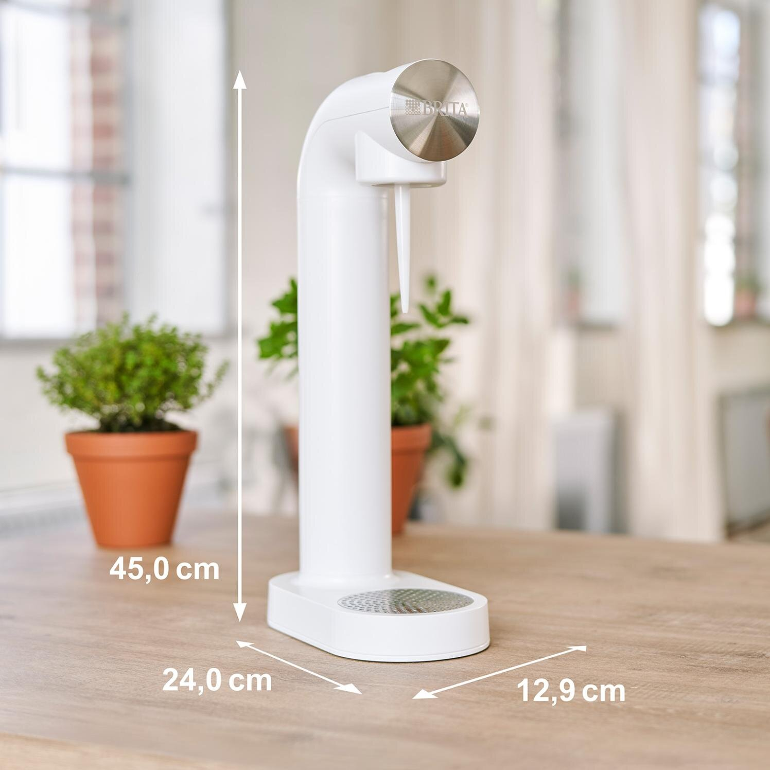 Biały dozownik wody z wymiarami. Na drewnianym stole, z roślinami w doniczkach.