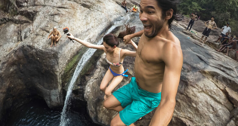 Mężczyzna i kobieta skaczą do wody ze skalistego klifu, inna osoba trzyma kamerę.