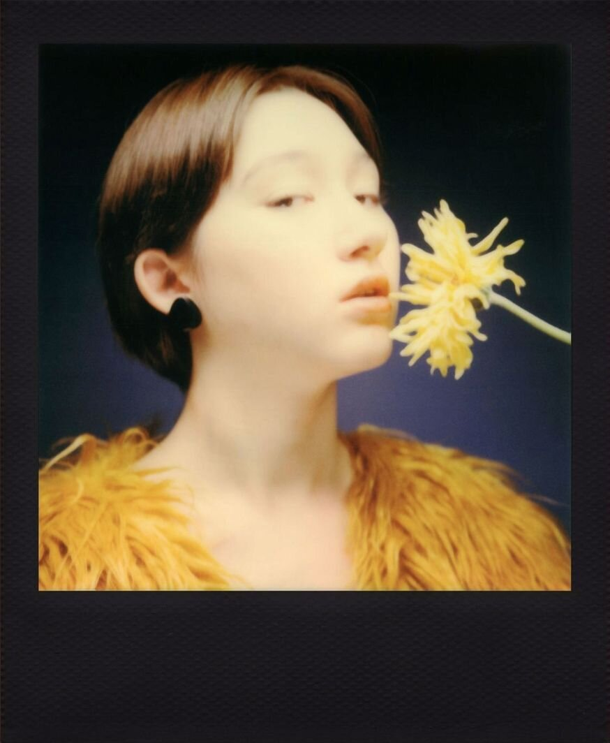 Kobieta z żółtym kwiatem, czarnymi kolczykami i futrem. Ciemne tło, ramka polaroid.