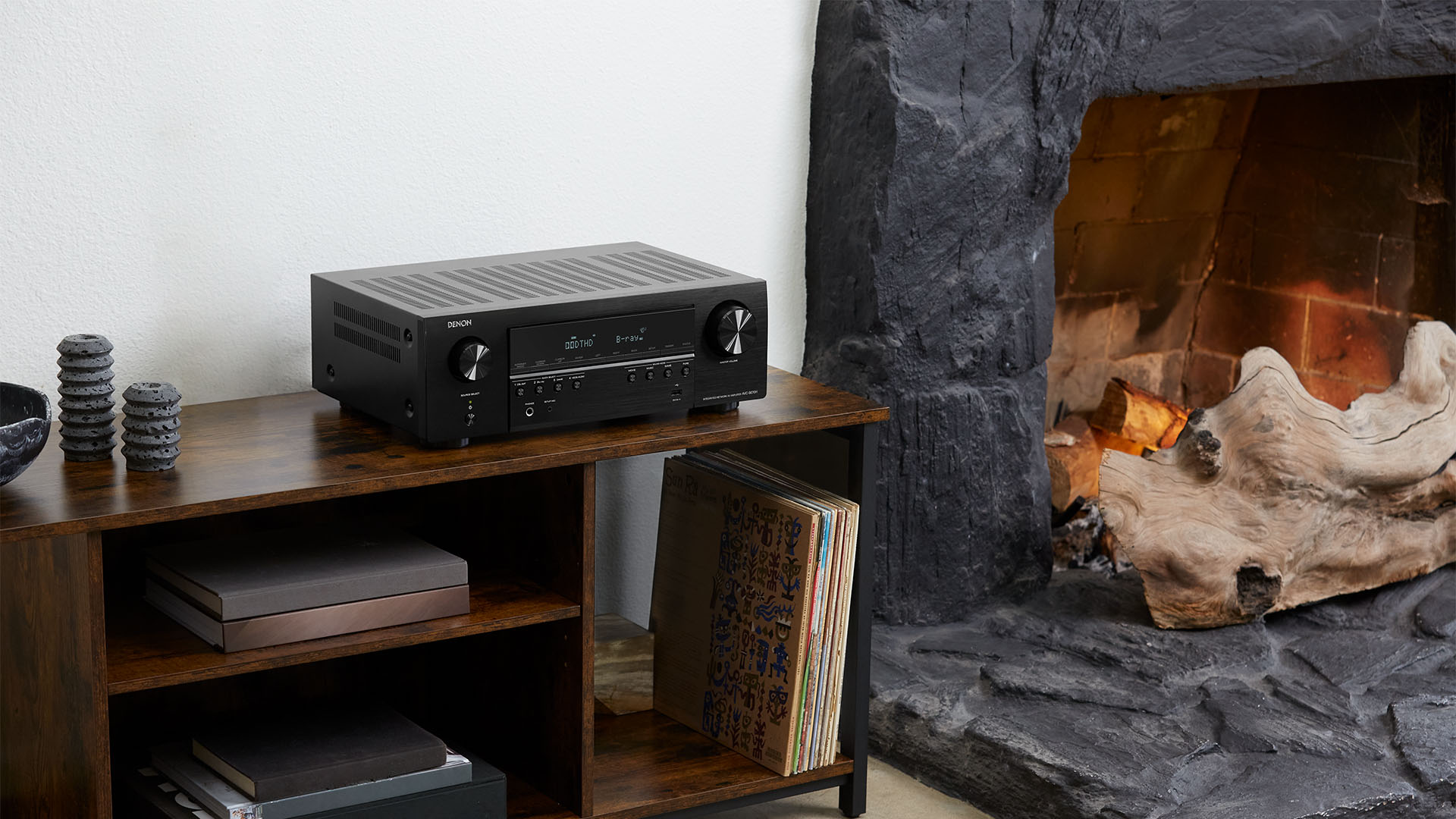 Ein schwarzer Denon-Receiver auf einem Holzregal. Ein Kamin ist im Hintergrund.