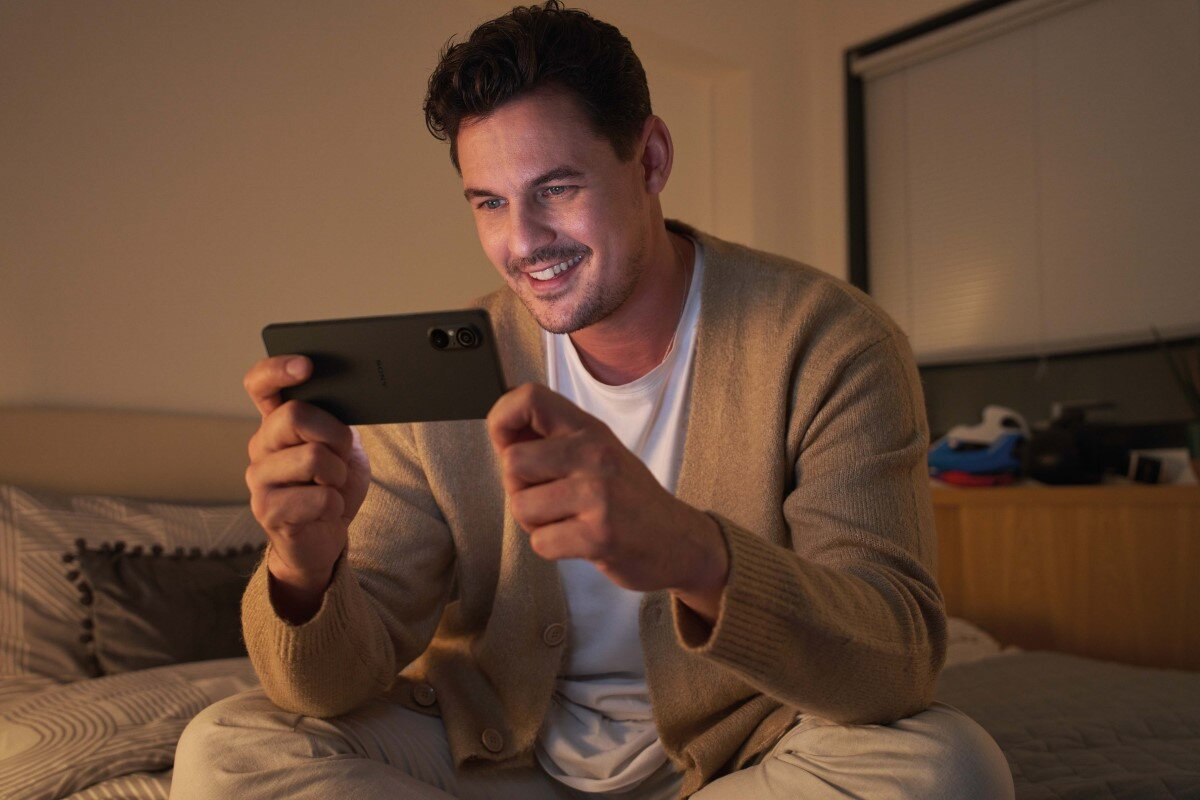 Mężczyzna w beżowym kardiganie siedzi na łóżku, trzymając czarny telefon.