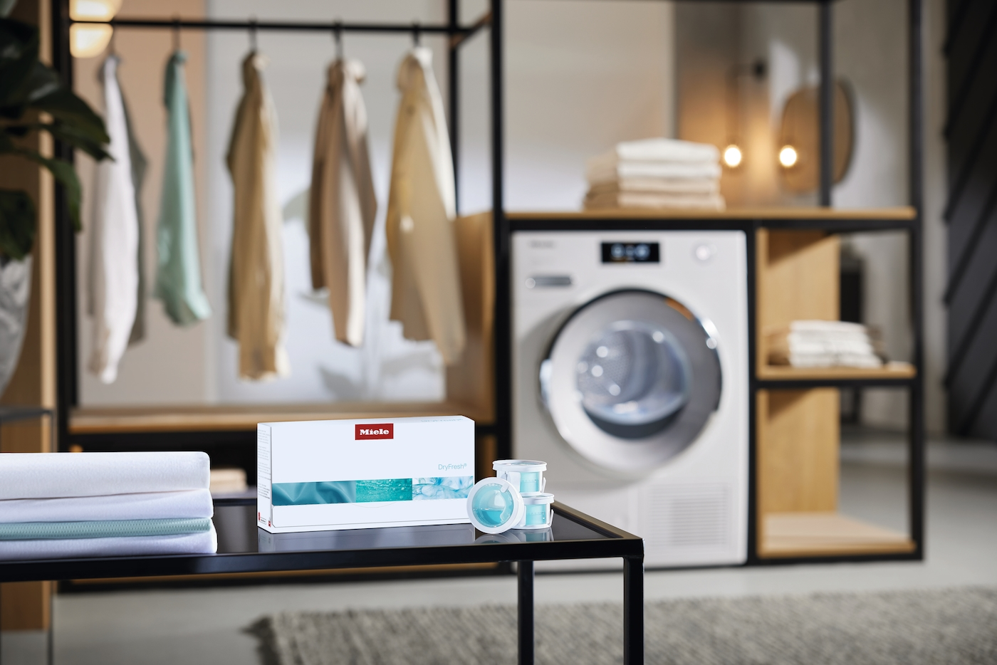 Ein Trockner steht neben einem Regal mit Kleidung. Daneben ein Tisch mit Miele-Produkten.