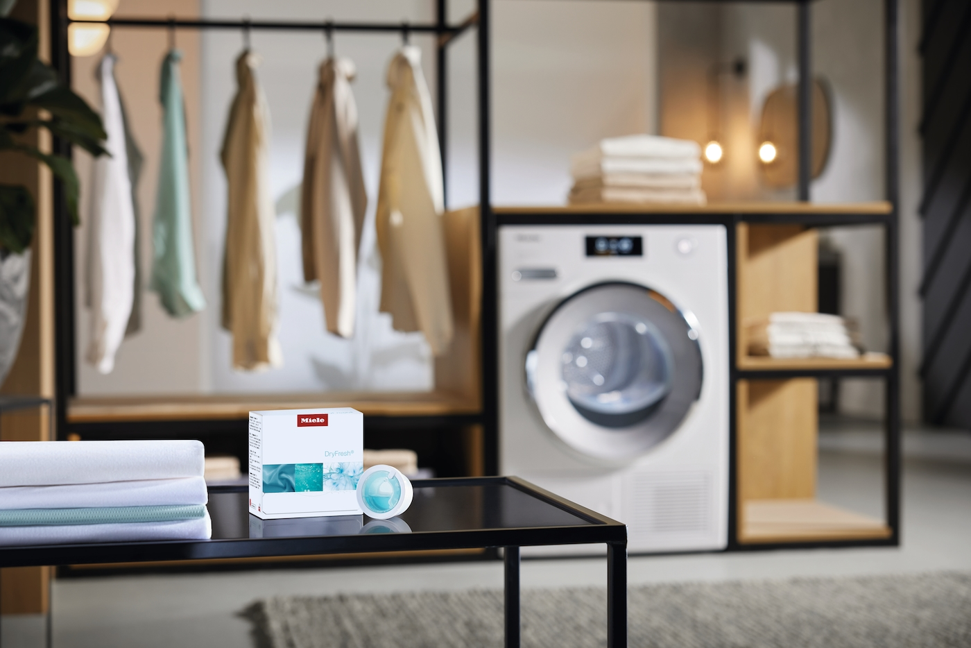 Ein Miele Trockner steht neben einem Regal mit Kleidung. Ein Tisch zeigt Wäsche und ein Produkt.