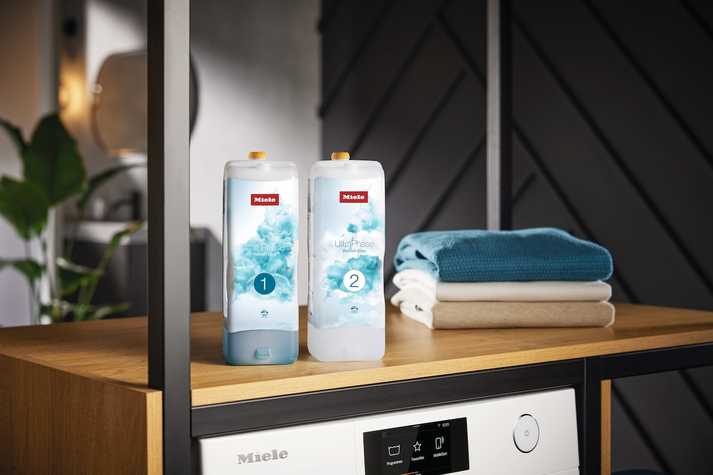 Zwei Miele UltraPhase Waschmittelflaschen, nummeriert 1 und 2. Daneben gefaltete Handtücher. Darunter eine Waschmaschine.
