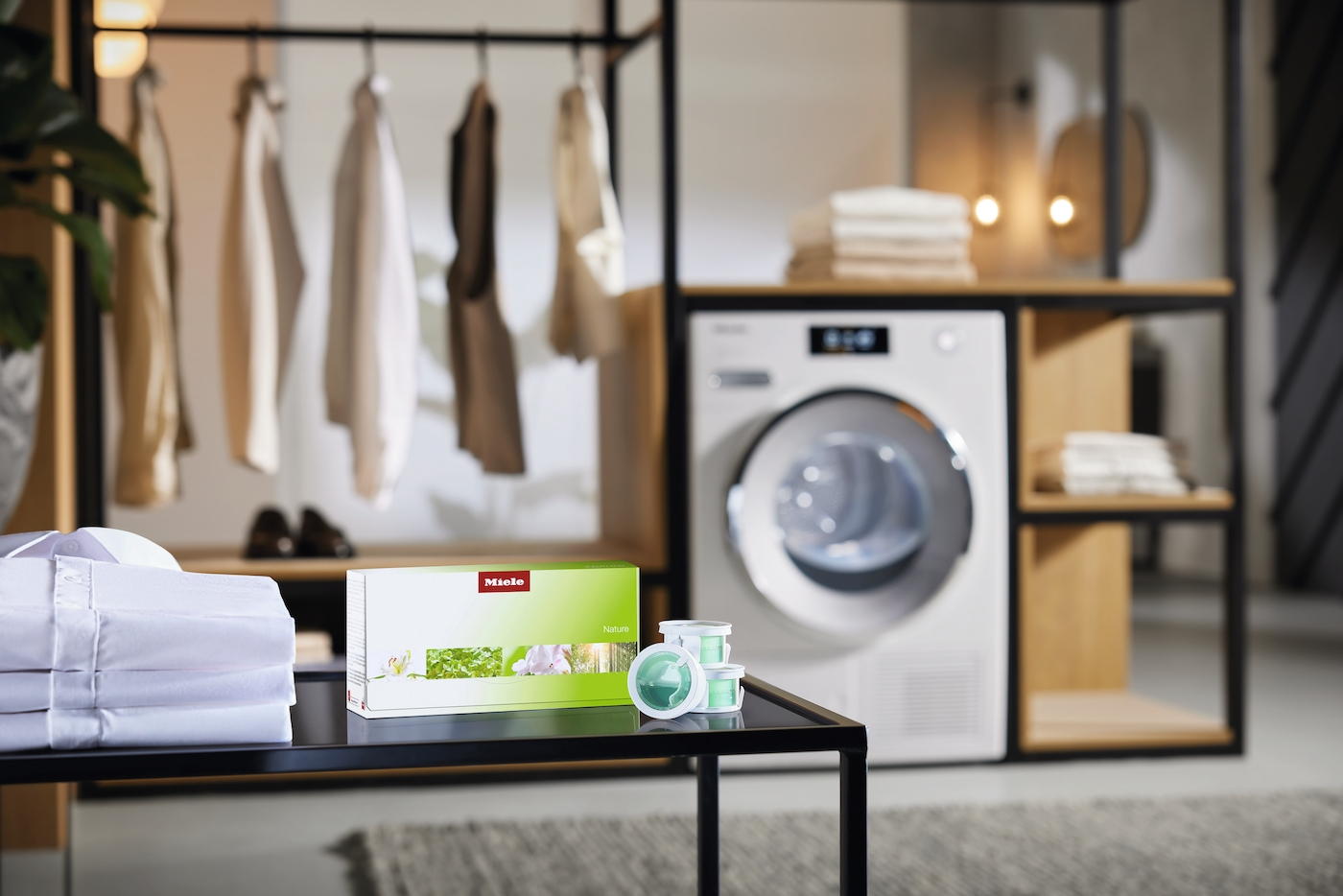 Ein Miele-Waschmittelpaket und Kapseln stehen auf einem Tisch vor einer Waschmaschine und Kleidung.