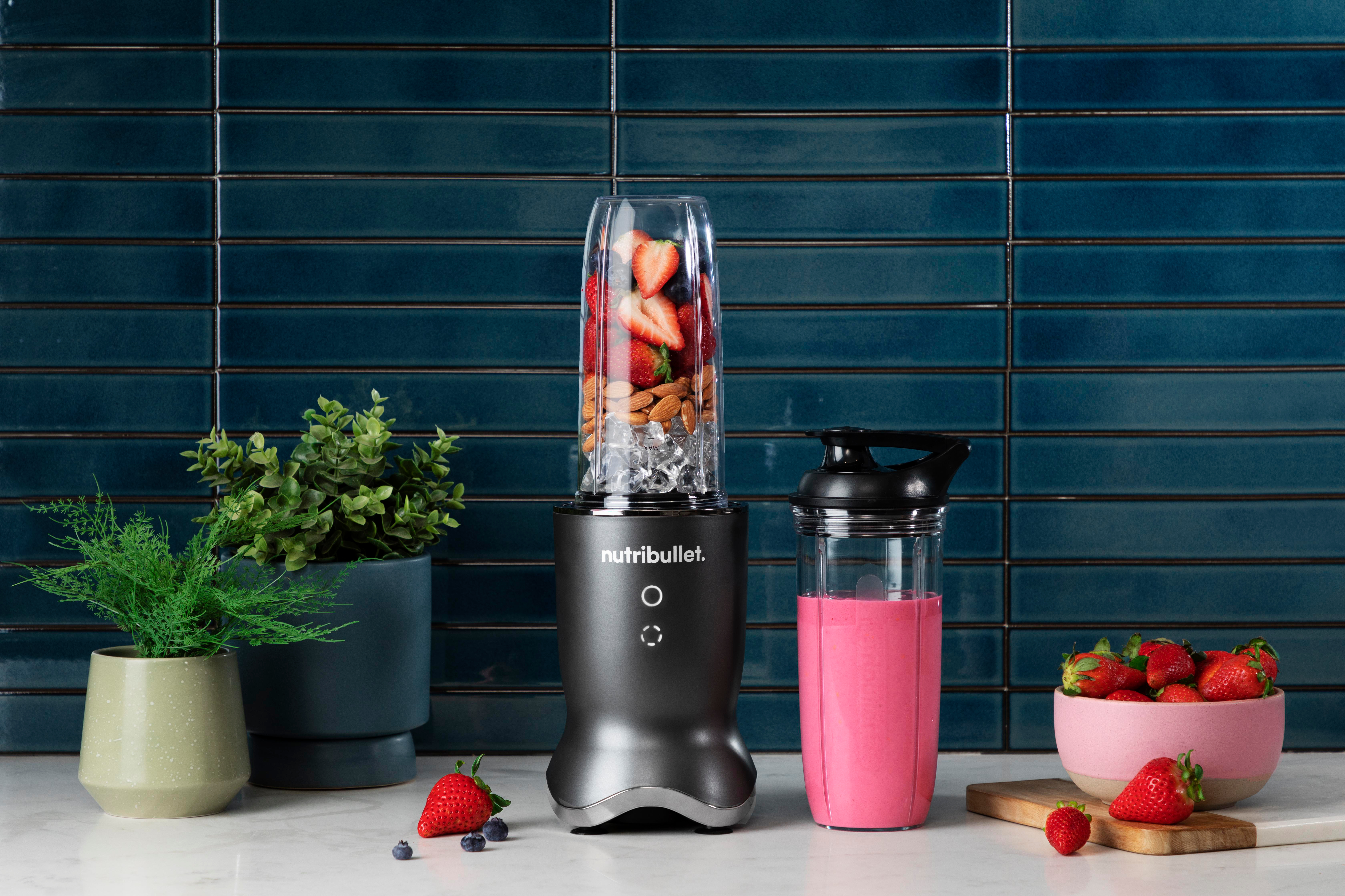 Mixer mit Früchten und Eis. Ein rosa Smoothie im Glas. Grüne Pflanze und Erdbeeren daneben.