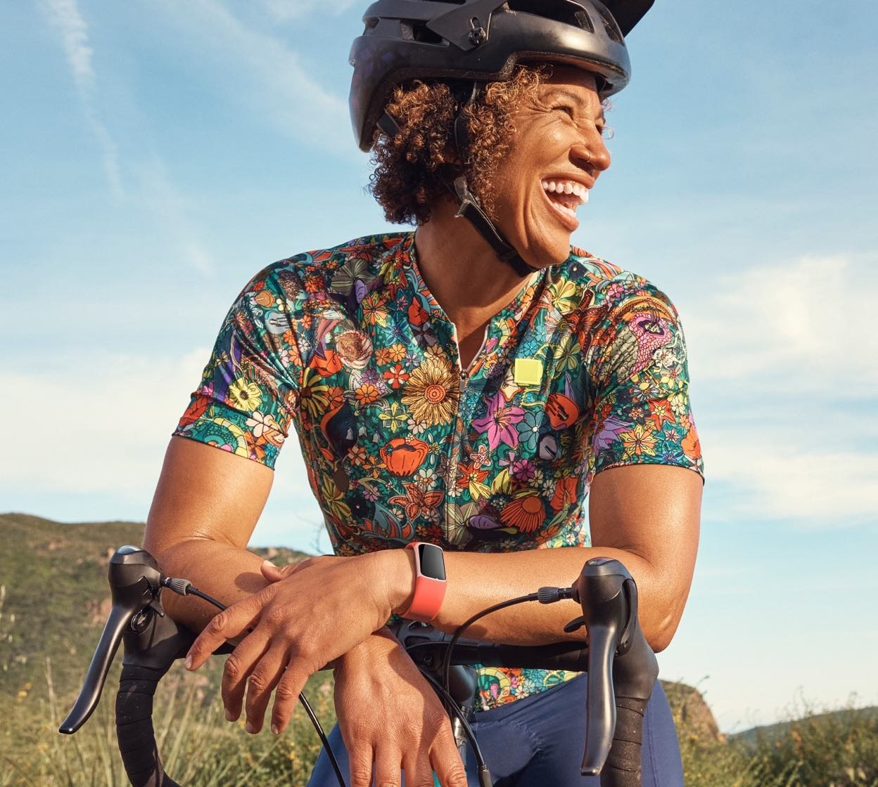 Ciclista sorridente, con una maglia floreale. Si appoggia alla sua bici, con un orologio al polso.