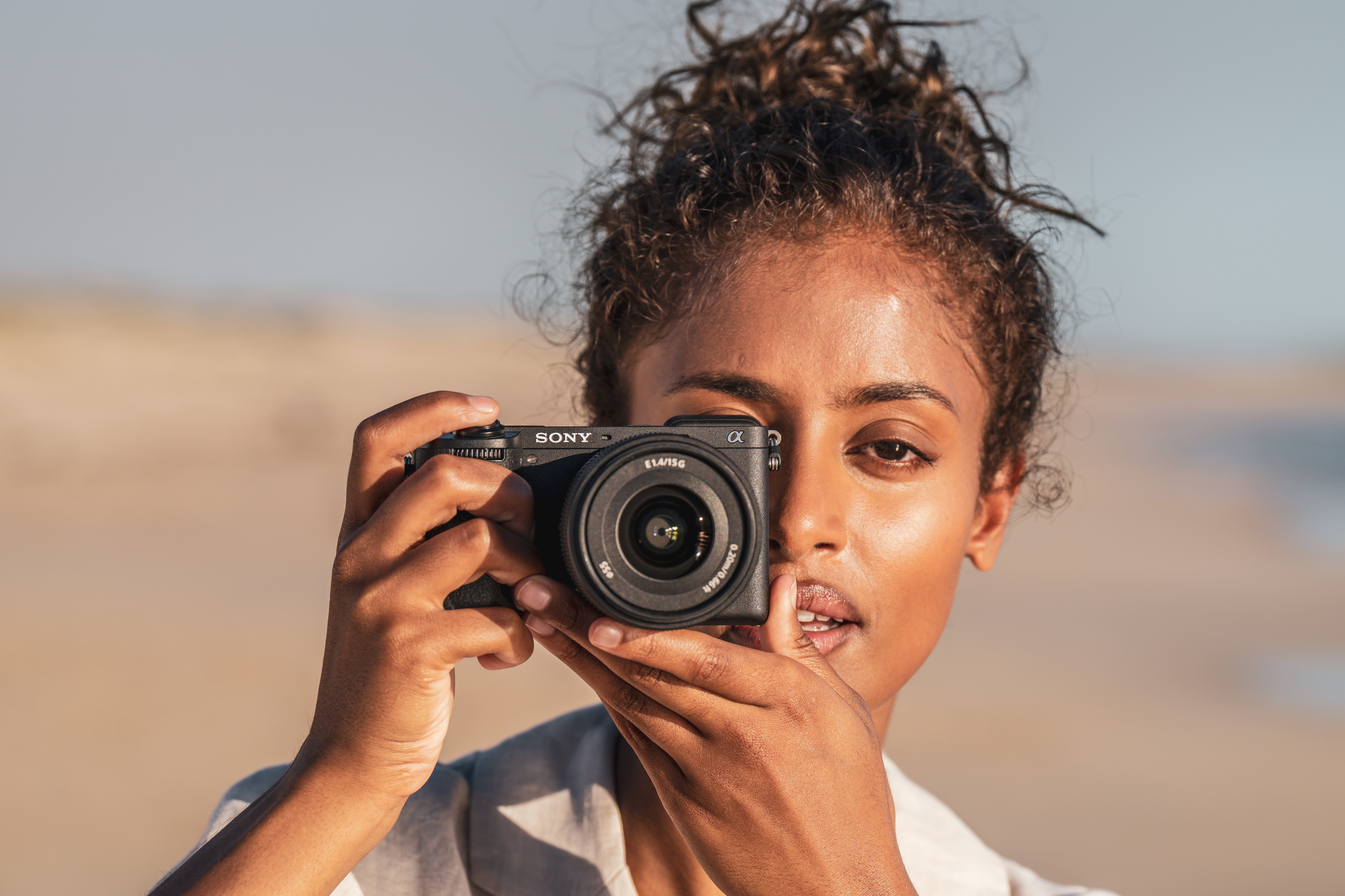 Una donna tiene in mano una fotocamera Sony nera, scattando una foto. Lo sfondo è una spiaggia soleggiata.