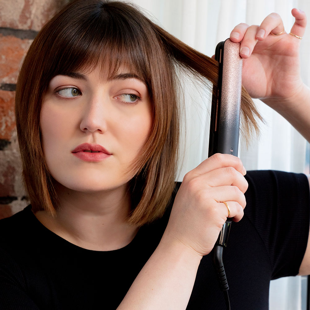Nő hajsimítót használ. A haját nézi. Téglatfal a háttérben.