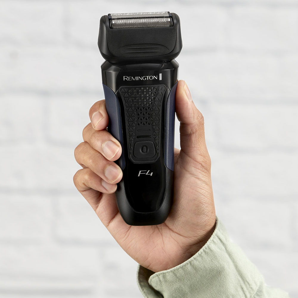 Egy kéz egy fekete-kék Remington F4 elektromos borotvát tart. Fehér tégla háttér.