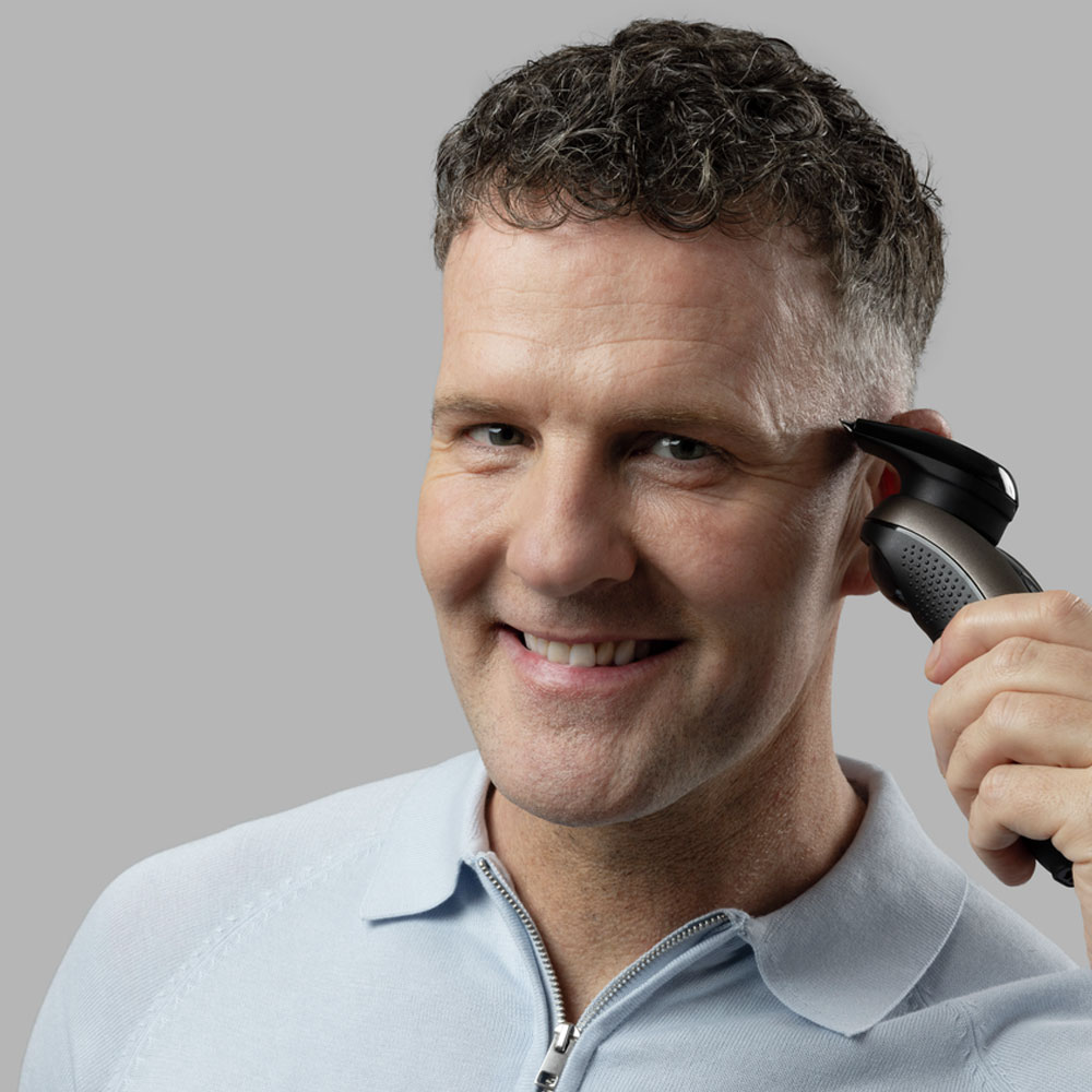Egy férfi elektromos borotvával borotválja a pajeszát, és a kamerába mosolyog.