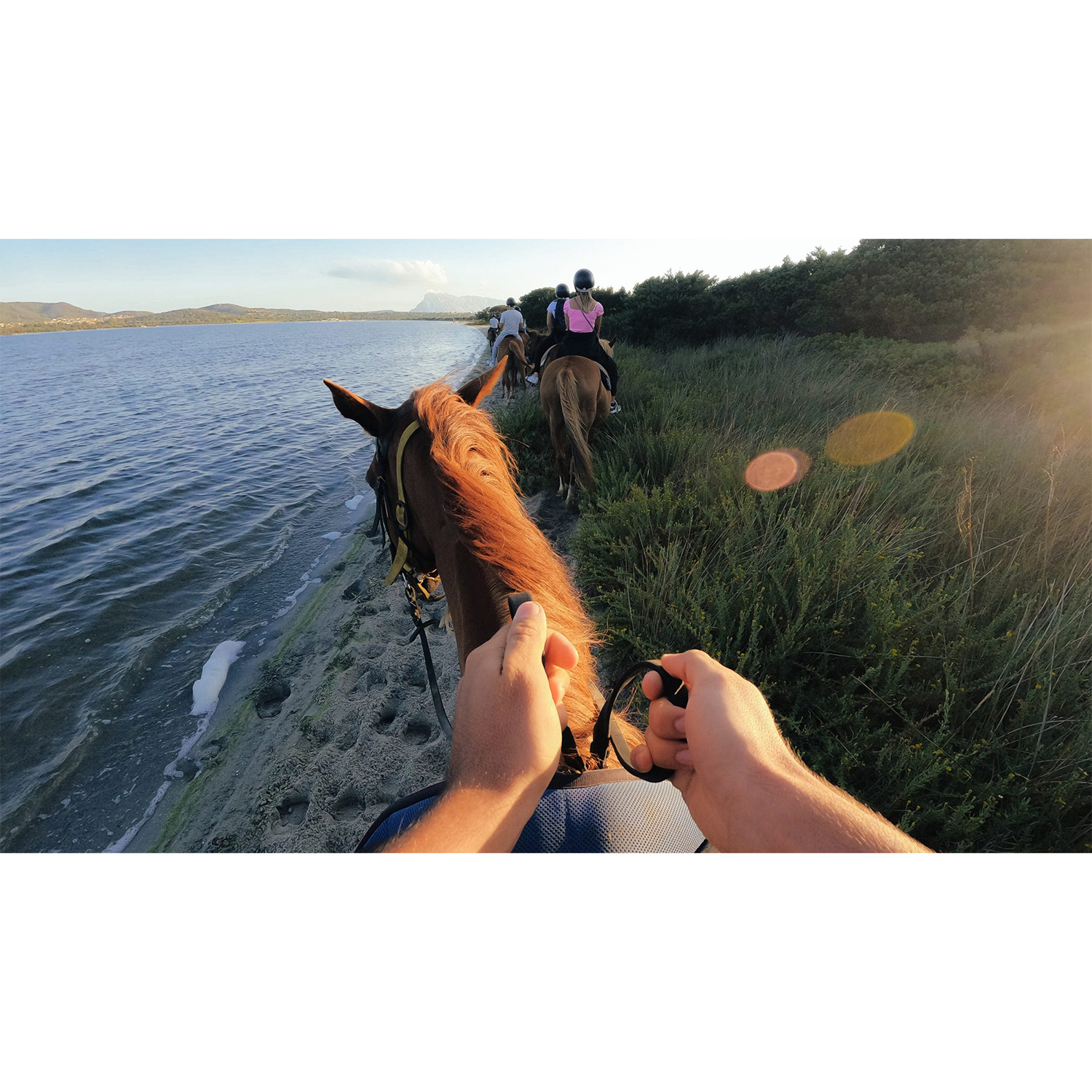 Persone a cavallo lungo la riva. Il sole tramonta, proiettando un bagliore caldo.