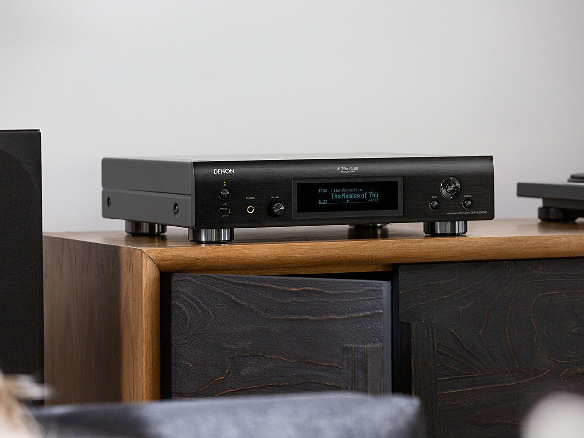 Ein schwarzes Audiogerät auf einem Holzschrank, mit einem Display, das Songinformationen anzeigt.