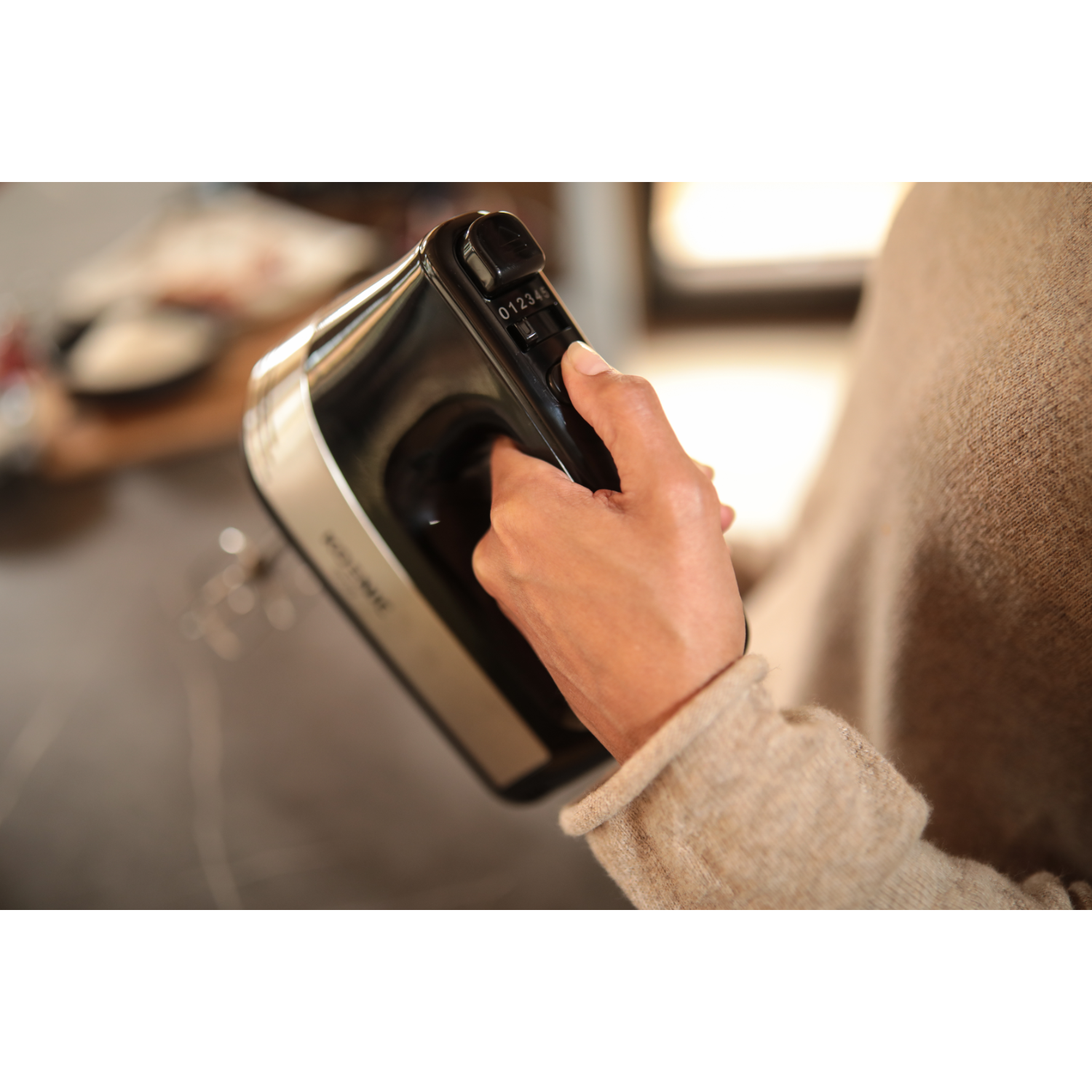 Hand holding a black and silver electric mixer. Numbers 0-4 visible. Beige sleeve.