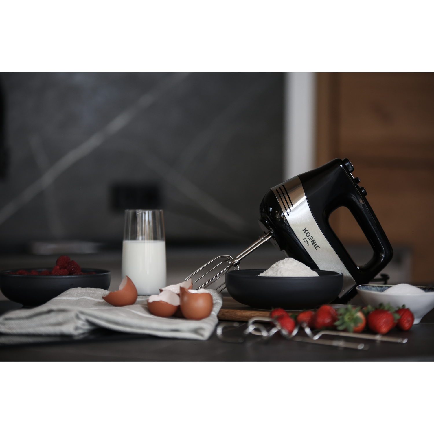 A mixer with ingredients like strawberries, milk, and eggs, on a table.