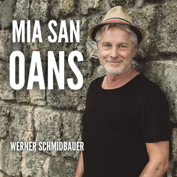 Mann in schwarzem Hemd und Hut steht vor einer Steinmauer. Text: Mia San Oans.