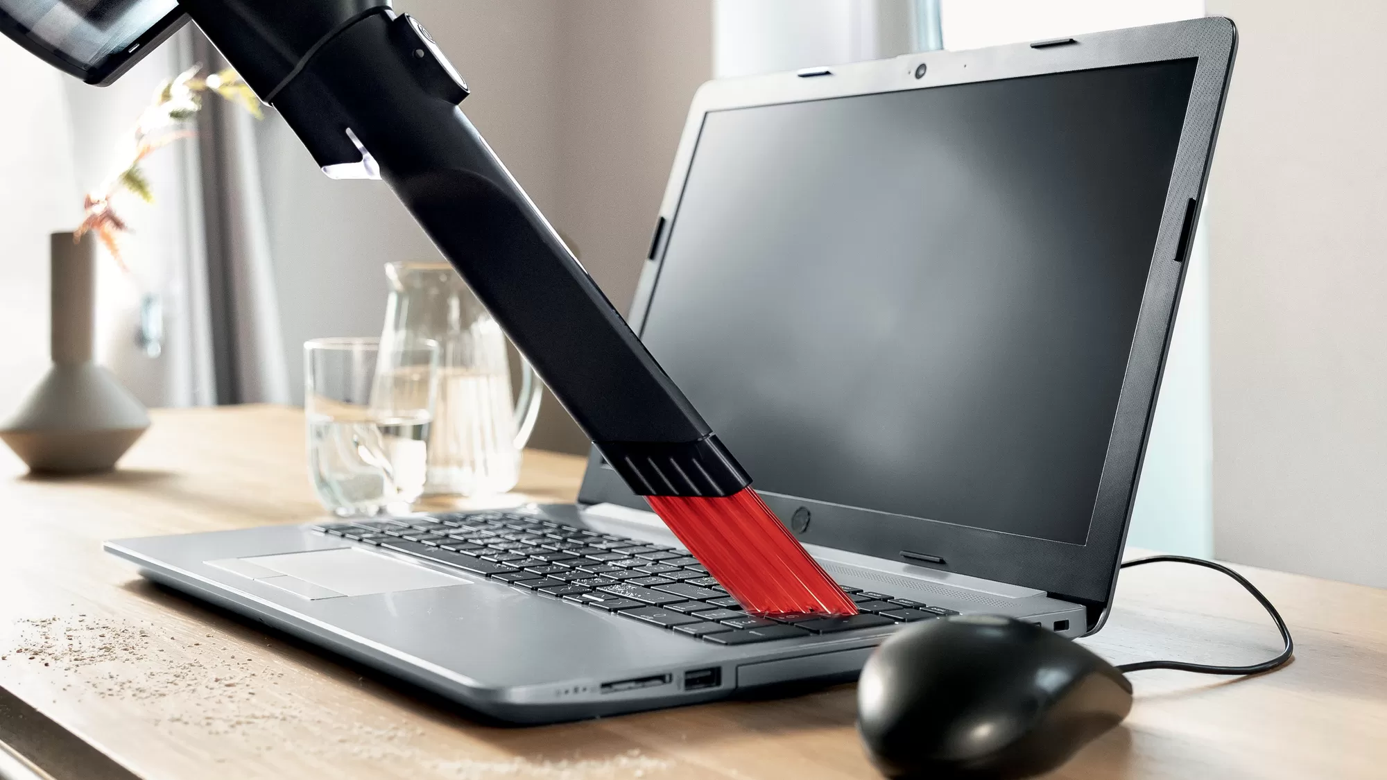 Egy fekete porszívófej tisztítja a laptop billentyűzetét. A képernyő fekete. Egy egér van az oldalon. Egy pohár víz van a háttérben.