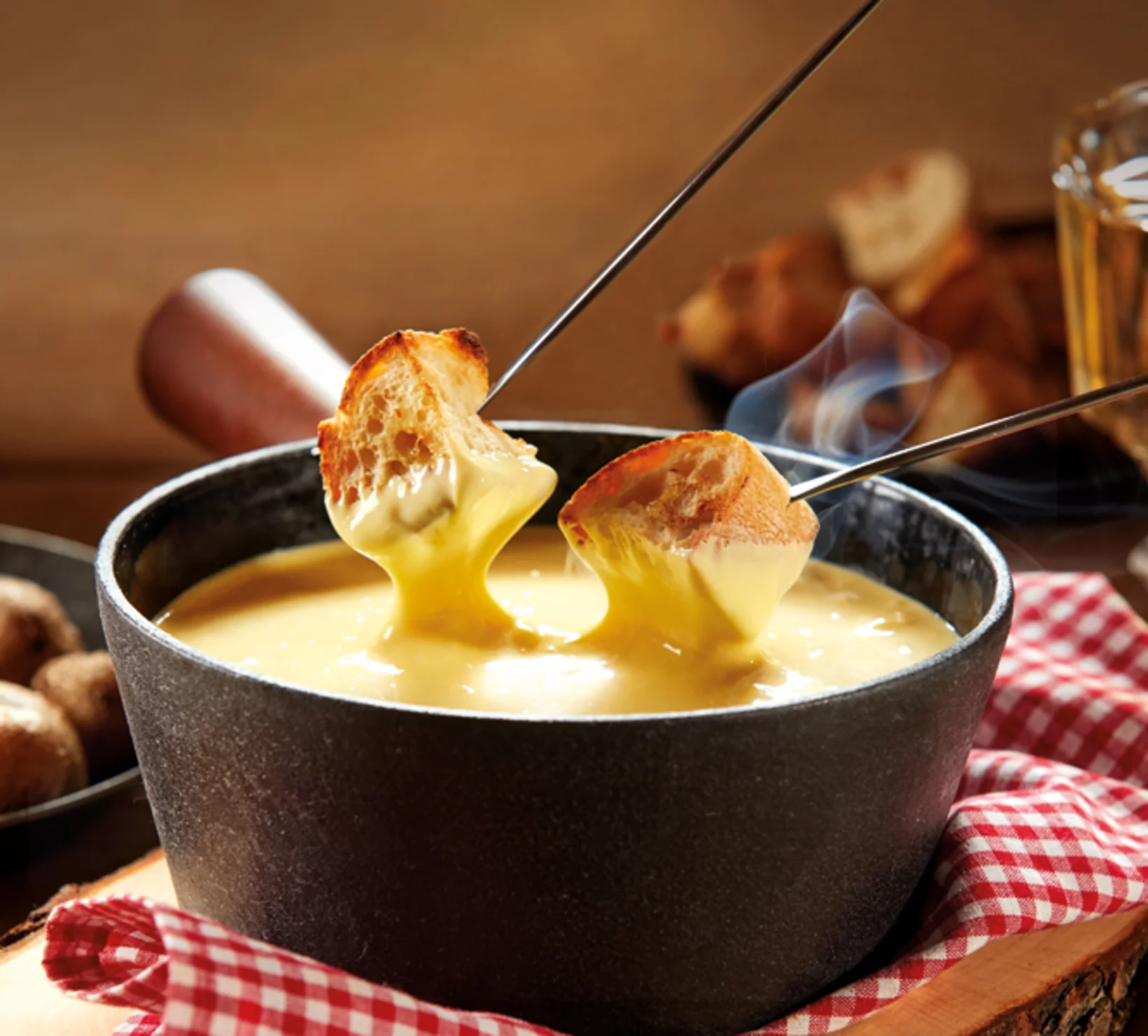 Fonduta con pane intinto nel formaggio fuso. Panno a scacchi rosso e bianco sotto. Sfondo in legno.