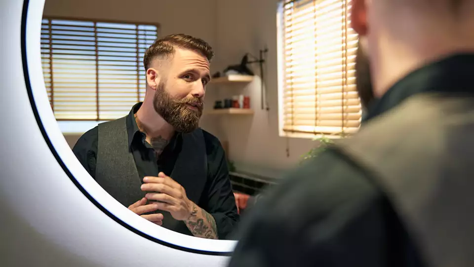 Homme se regardant dans un miroir, barbe stylisée, portant gilet et chemise, pièce bien éclairée.