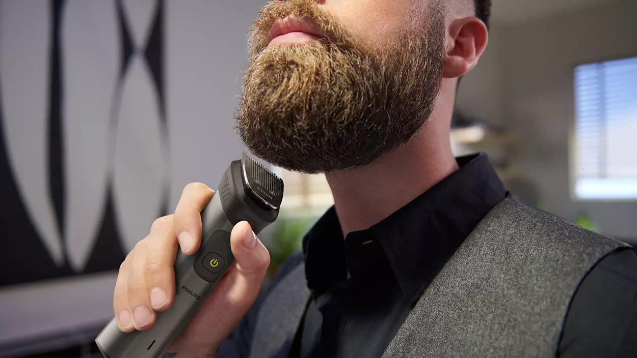 Un homme taille sa barbe avec une tondeuse à barbe Philips. La tondeuse est gris foncé et l'homme porte un gilet.