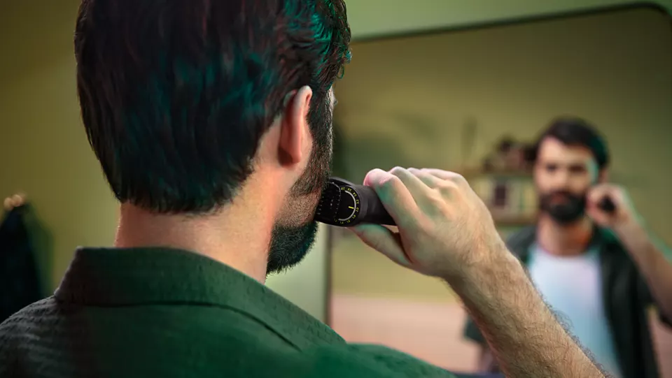 Un homme se rase la barbe avec une tondeuse, reflété dans le miroir. Mur vert.
