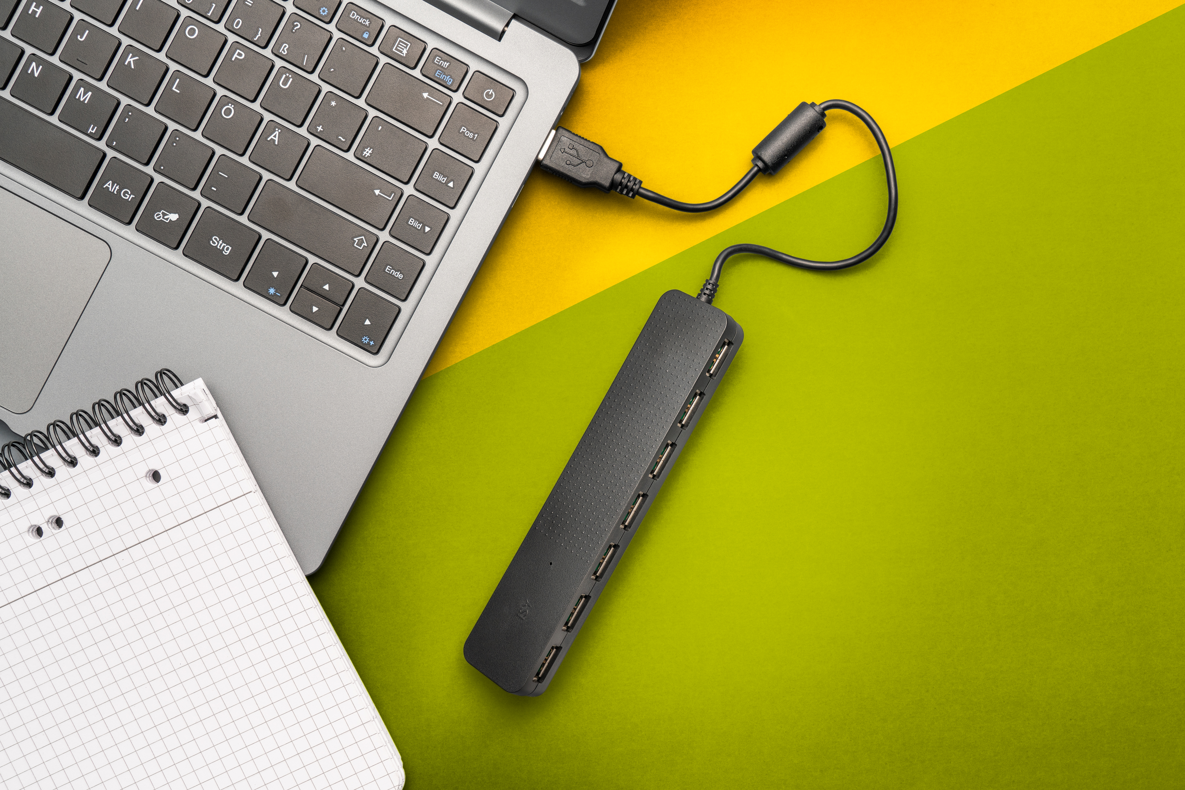 Ein Laptop, der mit einem USB-Hub verbunden ist, auf einer gelb-grünen Oberfläche mit einem Notizblock.