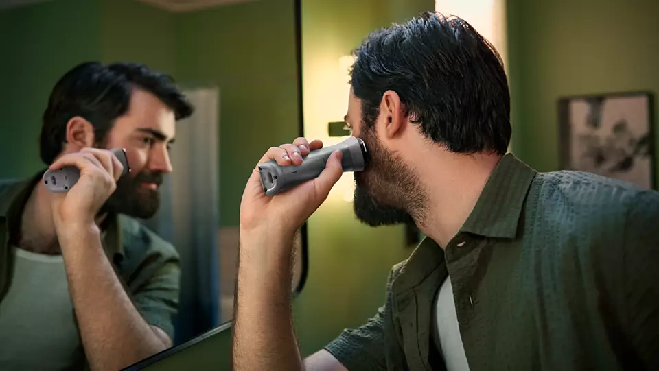 Uomo si rade la barba davanti a uno specchio con un rasoio elettrico grigio.