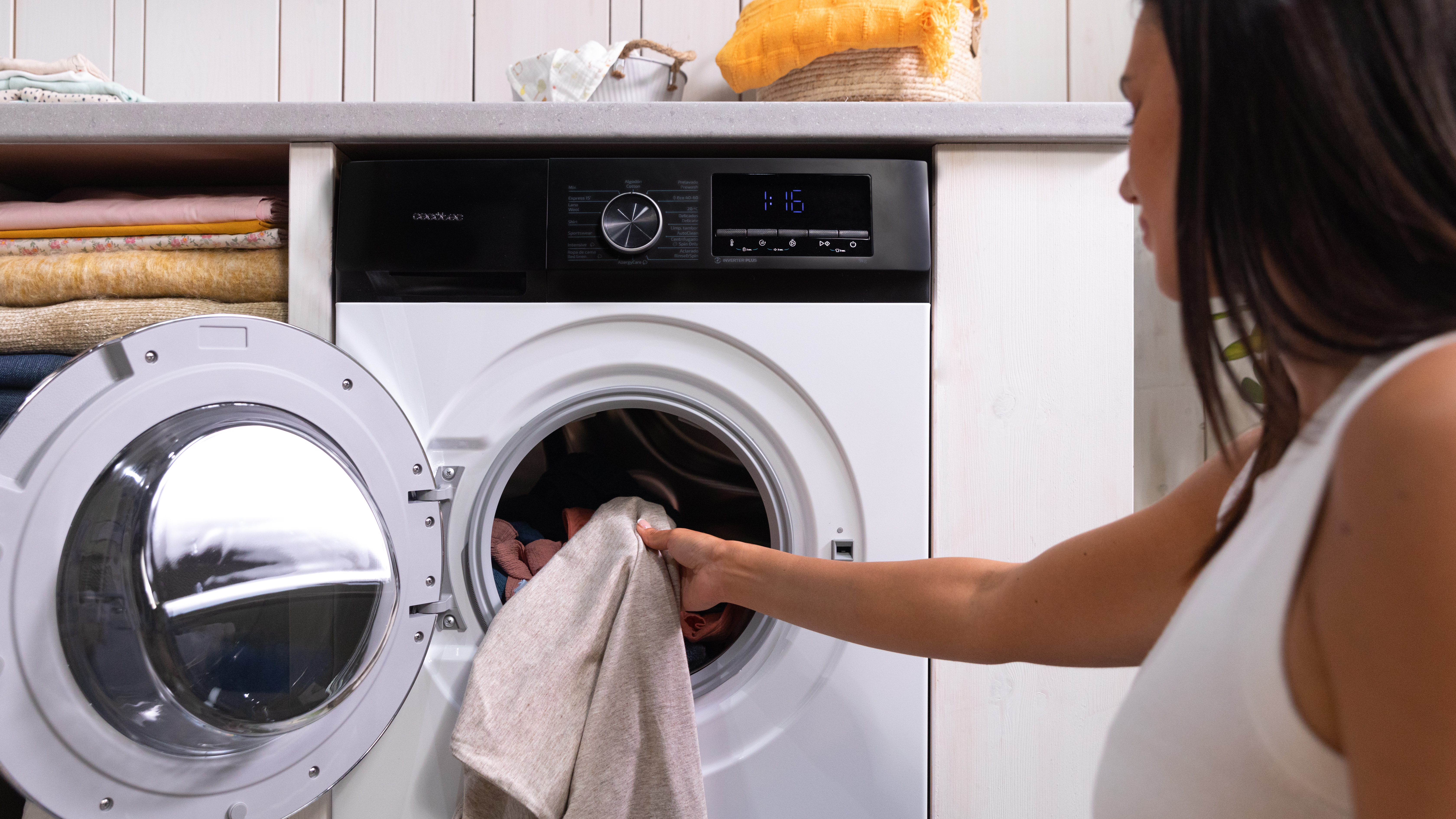 Mujer carga ropa en lavadora. Puerta abierta revela tambor. Otra ropa en estante sobre máquina.