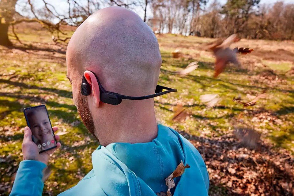 Mężczyzna w słuchawkach przewodnictwa kostnego patrzy na telefon. W tle drzewa i liście.