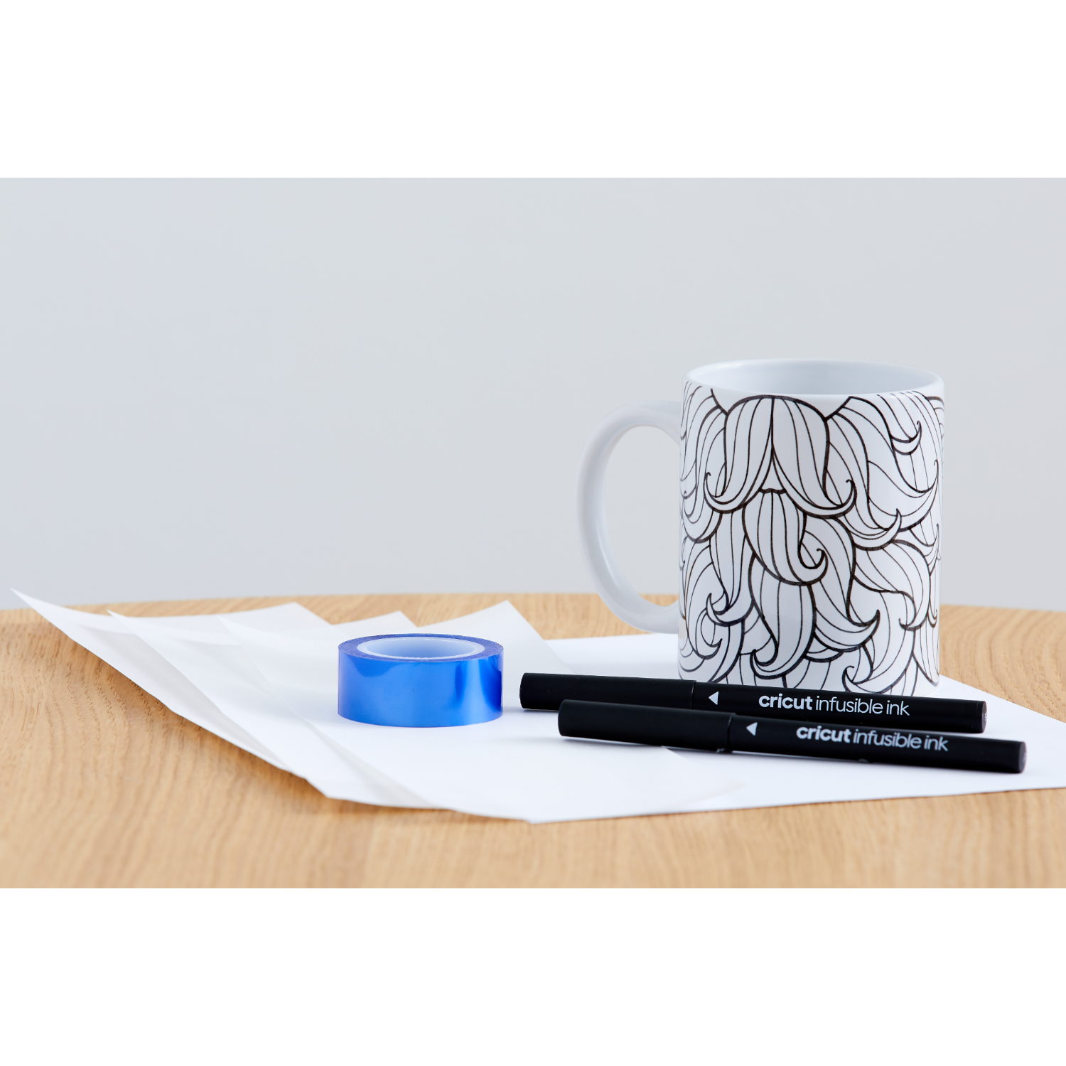Mug with black hair-like design, tape, and Cricut pens on wooden table.