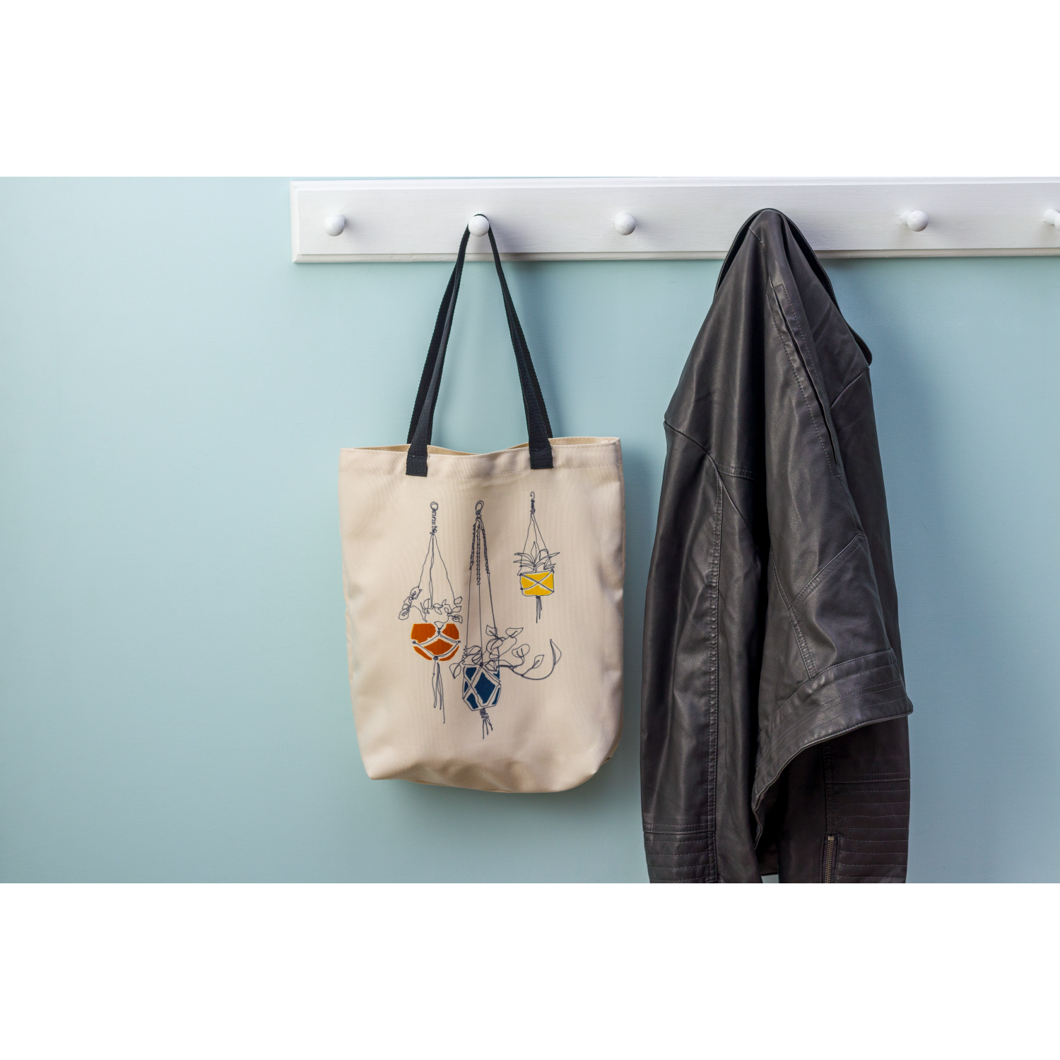 A tote bag with plant illustrations hanging on a wall with a jacket.