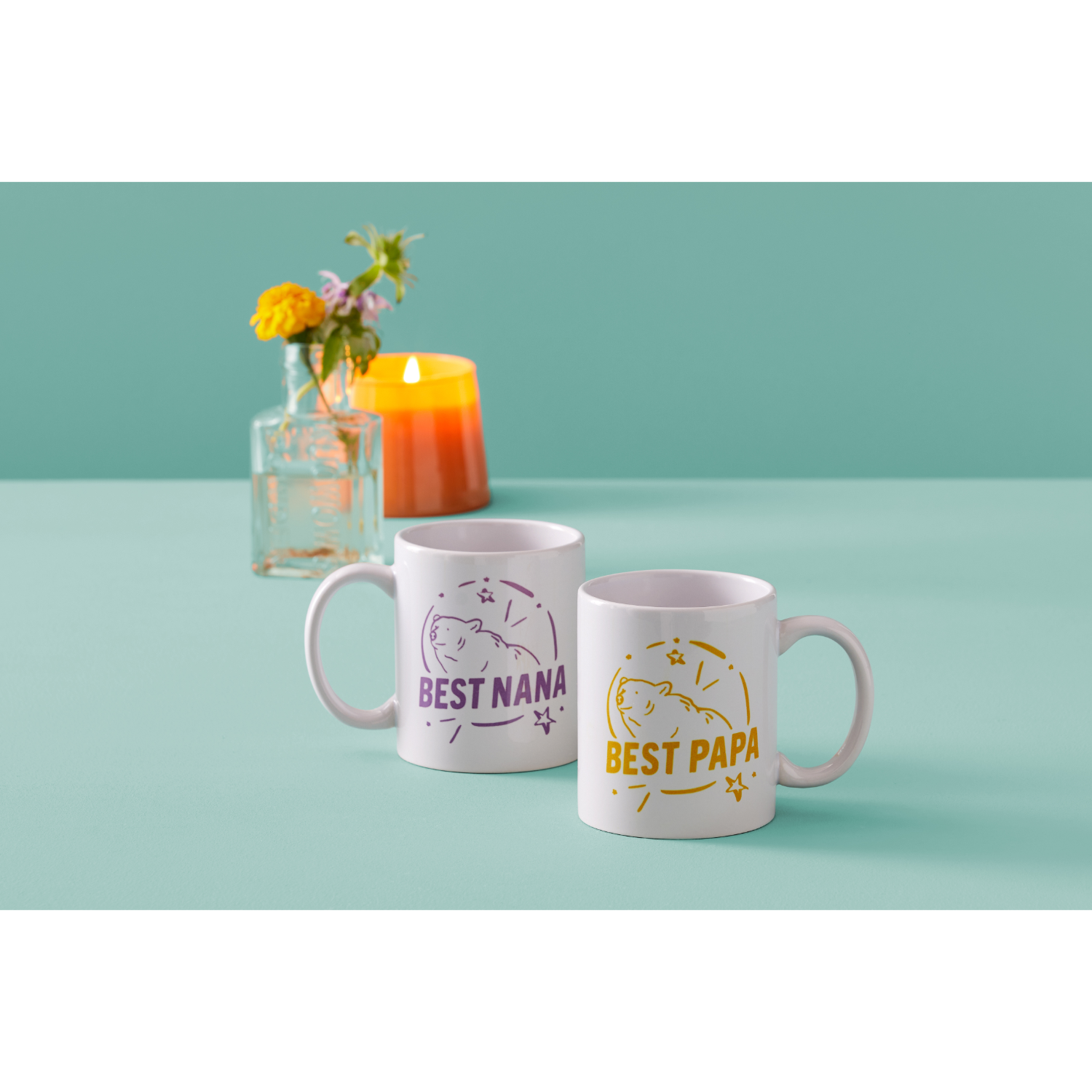 Two white mugs with text and cartoon bear graphics, with a flower vase and candle.